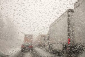 Cars and trucks driving on snow packed roads taken through a windshield covered with snowflakes.